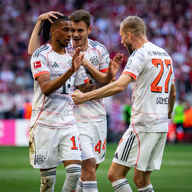 Three football players celebrate a goal on the field. One player raises his hands in excitement, while another places a hand on his head, showing camaraderie. They wear Bayern Munich's white and red team jerseys, with the stadium crowd blurred in the background.