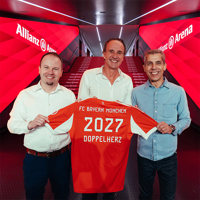 Three men stand together at the Allianz Arena, each holding a red football jersey featuring "FC Bayern München" and the number "2027 DOPPELHERTZ." They appear to be celebrating a significant announcement or partnership. The background shows a bright, modern corridor.