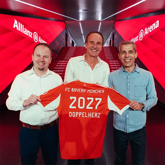 Three men stand together at the Allianz Arena, each holding a red football jersey featuring "FC Bayern München" and the number "2027 DOPPELHERTZ." They appear to be celebrating a significant announcement or partnership. The background shows a bright, modern corridor.