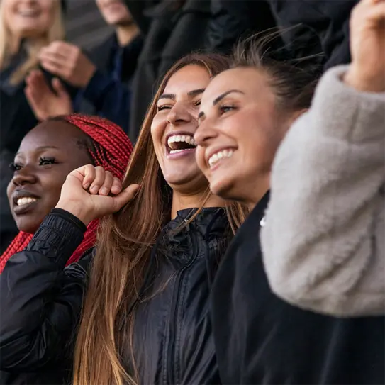 A group of three women are smiling and celebrating together, expressing joy and excitement. They are dressed in casual jackets, and their expressions convey happiness as they engage with their surroundings, likely at a lively event or gathering.